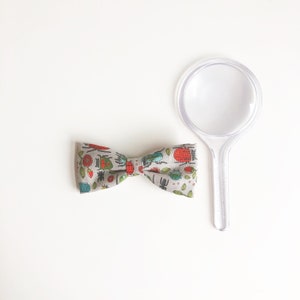 May include: A patterned bow tie featuring insects in red, green, and blue on a grey background, alongside a clear magnifying glass. The bow tie is made of fabric and has a traditional bow shape. The magnifying glass is clear plastic.
