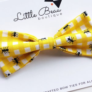 May include: A yellow and white gingham bow tie featuring a repeating pattern of black ants. The bow tie is on a white card with the text "Little Beau boutique" and "CRAFTED BOW TIES FOR ALL AGES."