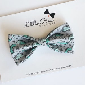 Puede incluir: Una pajarita gris con un estampado repetido de dinosaurios y palmeras verdes. La pajarita está colocada sobre una tarjeta blanca con el texto "Little Beau boutique" y una URL de la tienda Etsy.