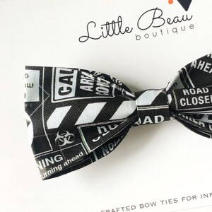 May include: A black and white fabric bow tie featuring a repeating pattern of various hazard warning signs, including biohazard symbols and text such as ROAD CLOSED, CAUTION, and WARNING, with the Little Beau boutique logo visible above.