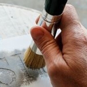 Puede incluir: Una mano sosteniendo un pincel con cerdas naturales, aplicando pintura gris a una plantilla sobre una superficie blanca. La plantilla dice "JUST".
