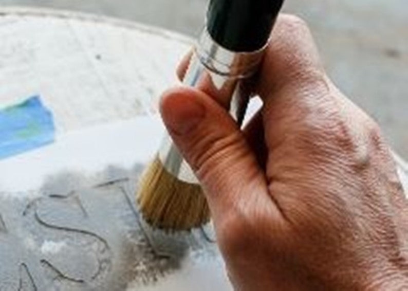 May include: A close-up of a hand holding a paint brush over a gray surface with the word 'JUST' written on it.