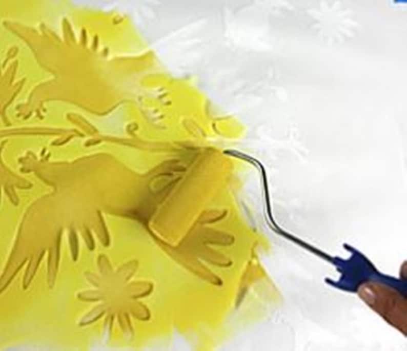 May include: A yellow paint roller with a blue handle is being used to paint a stencil of two birds and a flower on a white surface.