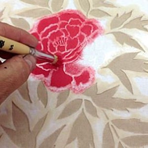 May include: A close-up of a hand using a paintbrush to apply pink paint through a stencil onto a white surface. The stencil is a floral design with a large flower in the center and smaller leaves surrounding it. The paintbrush is made of natural bristles and has a wooden handle.
