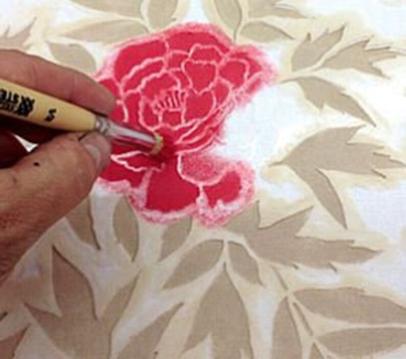 May include: A close-up of a hand using a paintbrush to apply pink paint through a stencil onto a white surface. The stencil is a floral design with a large flower in the center and smaller leaves surrounding it. The paintbrush is made of natural bristles and has a wooden handle.