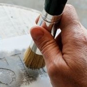 May include: A hand holding a paintbrush with natural bristles, applying gray paint to a stencil on a white surface. The stencil reads "JUST".