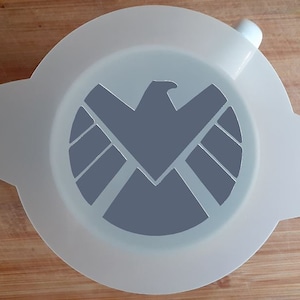 Puede incluir: Plantilla de plástico blanco con un recorte gris del logotipo de S.H.I.E.L.D., que presenta un halcón con las alas extendidas y un escudo circular con una estrella en el centro.