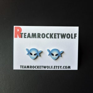 May include: A pair of blue and white earrings featuring a stylized face with wings. The earrings are on a white card with the text "TEAMROCKETWOLF.ETSY.COM".