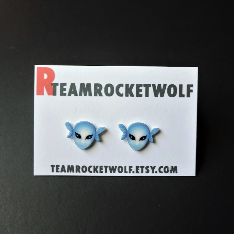 May include: A pair of blue and white earrings featuring a stylized face with wings. The earrings are on a white card with the text "TEAMROCKETWOLF.ETSY.COM".