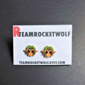 May include: A pair of earrings featuring a green-haired character with a red and blue mask. The earrings are on a white card with the text "TEAMROCKETWOLF.ETSY.COM".