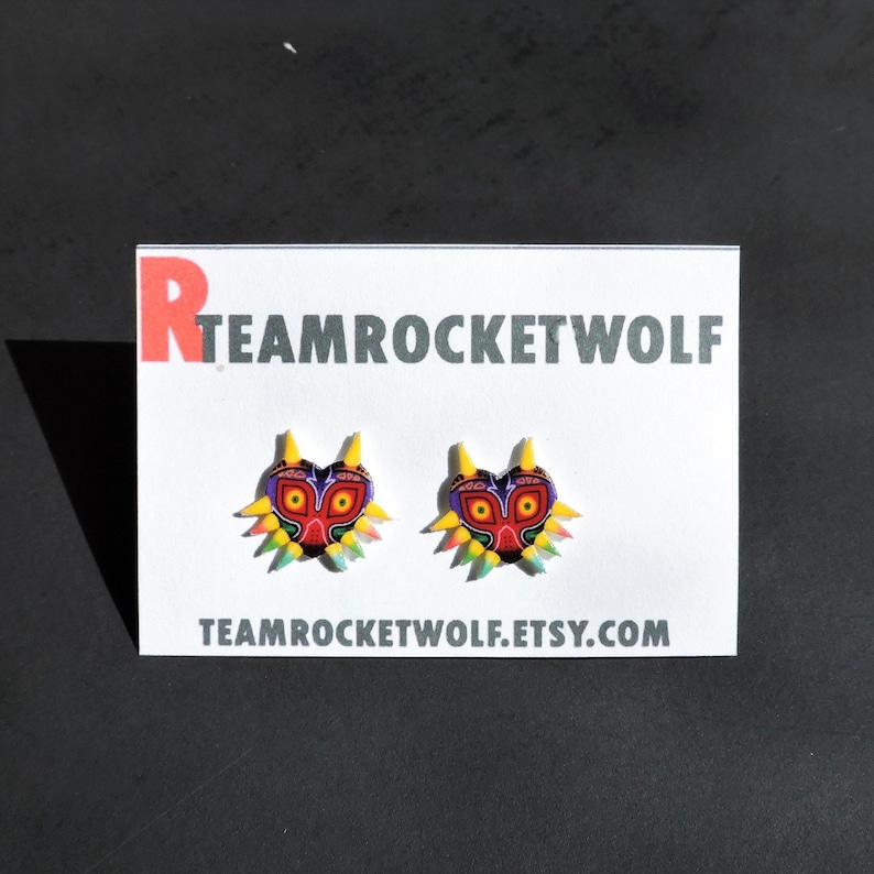 May include: A pair of earrings featuring a colorful Majora's Mask design. The earrings are on a white card with the text "TEAMROCKETWOLF.ETSY.COM".