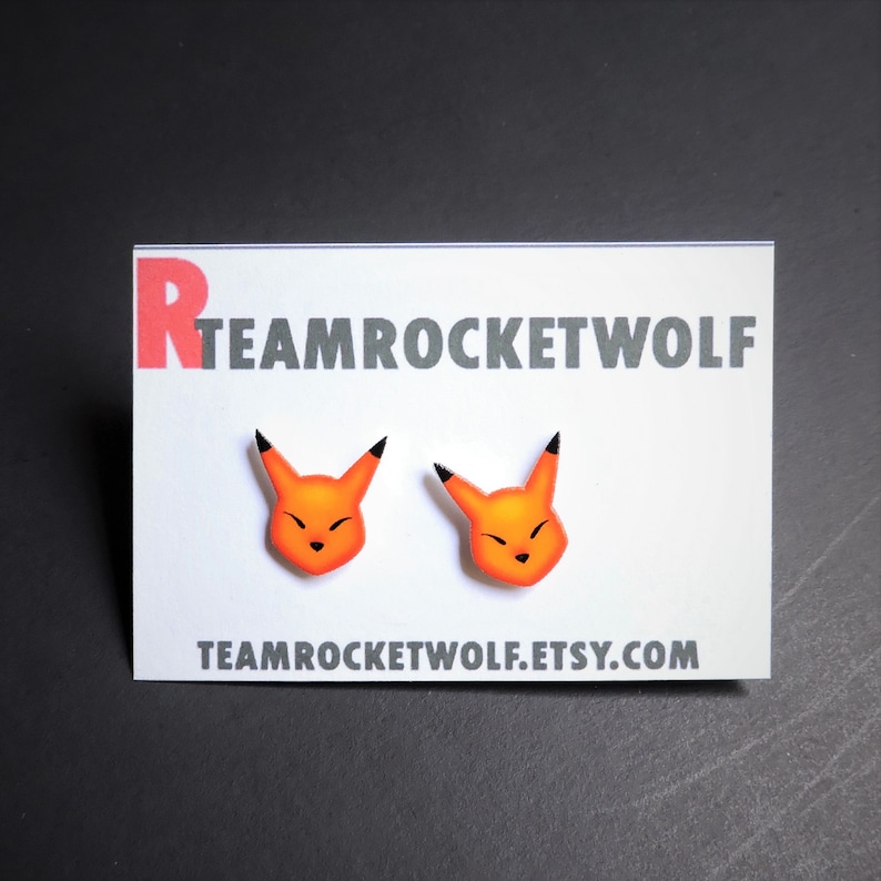 May include: A pair of orange and black cartoon fox earrings. The earrings are on a white card with the text "TEAMROCKETWOLF.ETSY.COM".