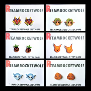 May include: Four sets of earrings featuring characters from the Legend of Zelda video game series. The first set features Majora's Mask, the second set features the character Sheik, the third set features the character Deku Scrub, and the fourth set features the character Goron.