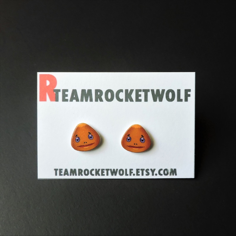 May include: A pair of orange and purple cartoon face earrings on a white card with the text "TEAMROCKETWOLF.ETSY.COM" and "TEAMROCKETWOLF" in red and black text.