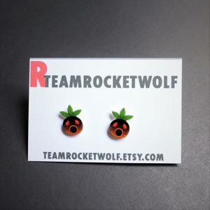 May include: A pair of earrings featuring a cartoon character with a red mouth and black eyes. The character is orange and brown with green leaves on top. The earrings are on a white card with the text "TEAMROCKETWOLF.ETSY.COM".