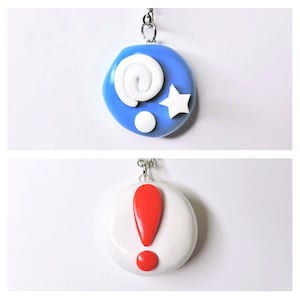 Puede incluir: Dos encantos hechos a mano de arcilla polimérica en cadenas plateadas. El primer encanto es azul con un remolino blanco, un punto blanco y una estrella blanca. El segundo encanto es blanco con un signo de exclamación rojo.