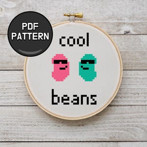 May include: A cross-stitch embroidery hoop featuring the words "cool beans" with pixelated bean characters wearing sunglasses. The design includes a pink and a teal bean. A black circle with "PDF PATTERN" is in the upper left.