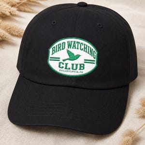 Op de afbeelding: Een zwarte honkbalpet met een geborduurde ovale patch op de voorkant. De patch heeft een witte achtergrond met een groene rand, groene tekst BIRD WATCHING CLUB, een groen vogelsilhouet en de tekst PHILADELPHIA, PA.