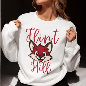May include: White sweatshirt featuring a red fox illustration with the words "Flint Hill" in red script. The fox has a smiling expression. The sweatshirt has long sleeves.