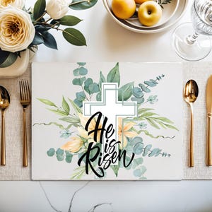 Op de afbeelding: Witte placemat met een aquarelontwerp van een kruis, groene bladeren en gele bloemen. De tekst "He is Risen" is geschreven in zwarte cursieve letters.