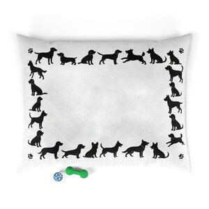Puede incluir: Cama para perros blanca con un borde de siluetas de perros negros y huellas de patas. Una pelota azul y blanca y un juguete verde con forma de hueso están en la parte inferior. La cama es rectangular y tiene un aspecto suave y afelpado.