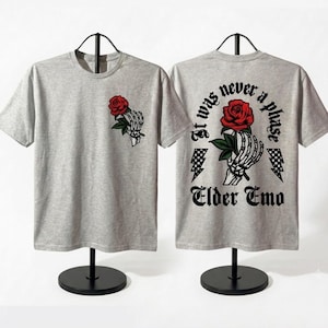 May include: A heather gray t-shirt featuring a skeleton hand holding a red rose on the front left chest. The back displays a larger skeleton hand holding a red rose, checkered lightning bolts, and the text: It was never a phase Elder Emo.