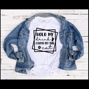 May include: White t-shirt with black text that reads "Hold my drink I gotta pet this cat" with a paw print and a drink icon.