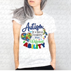 May include: White t-shirt featuring the word "Autism" in blue script. The shirt includes a colorful puzzle heart and the phrase "It's Not A Disability, It's A Different Ability" in various colors. A ribbon is also present.