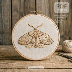 Puede incluir: Un bordado detallado de una polilla en tonos marrones, centrado en una tela beige dentro de un bastidor de bordado de madera. El texto "PDF PATTERN" es visible, junto con información adicional. El logotipo de The Crafty Studio está abajo.