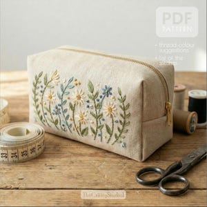 Könnte beinhalten: Eine beige rechteckige Kosmetiktasche mit floralem Stickdesign. Die Stickerei zeigt weiße, gelbe und blaue Blumen mit grünen Blättern. Die Tasche hat einen goldenen Reißverschluss und den Text "PDF PATTERN".