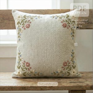 Può includere: Un cuscino quadrato in lino beige con disegni floreali ricamati agli angoli. Il ricamo presenta fiori bianchi, rosa e blu con foglie verdi. Il cuscino è su una panca di legno. Il testo sul cuscino recita "PDF PATTERN".