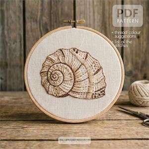 Op de afbeelding: Geborduurd zeeschelpontwerp in een houten hoepel. De beige stof heeft een gedetailleerd bruin zeeschelppatroon. De tekst "PDF PATTERN" is zichtbaar, samen met extra tekst. Het logo van The Crafty Studio staat onderaan.