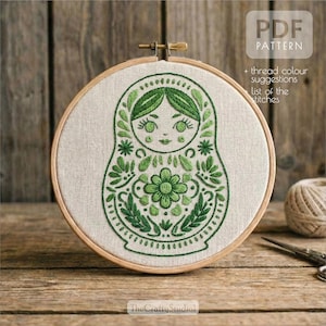 May include: An embroidered Matryoshka doll design in a wooden hoop, featuring green thread. The design includes floral and leaf patterns, with the text "PDF PATTERN" and "The Crafty Studio" visible.
