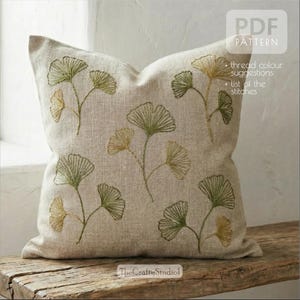 May include: A square beige cushion featuring embroidered ginkgo leaf designs in green and gold. The cushion is a PDF pattern with thread colour suggestions and a list of the stitches. The Crafty Studio logo is visible.
