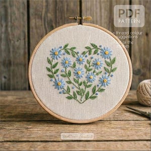 Könnte beinhalten: Ein Stickmuster in Herzform mit blauen Blumen und grünen Blättern, in einem Holzrahmen. Der Text "PDF PATTERN" ist sichtbar. Das Logo The Crafty Studio befindet sich unten.