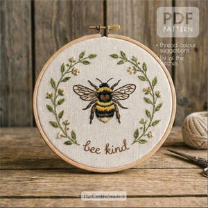 May include: Embroidered bee design in a wooden hoop. The bee is black and yellow, surrounded by green leaves and small white flowers. The words "bee kind" are stitched below the bee. The text "PDF PATTERN" is in the upper right corner.
