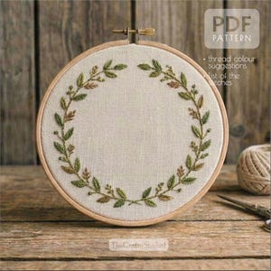 May include: A round embroidery hoop with a cream-colored fabric featuring a green and brown leaf design. The text "PDF PATTERN" is visible, along with additional text. The hoop is set on a wooden surface, with a ball of thread and scissors nearby.