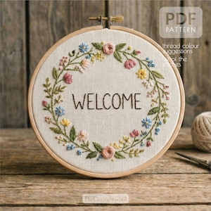 May include: Embroidered hoop art with the word "WELCOME" in brown thread, surrounded by a floral wreath of pink, blue, and yellow flowers. The design is set against a beige fabric background, held in a wooden hoop.