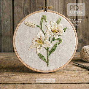 以下が含まれることがあります： 木製の刺繍枠に刺繍されたユリの花のデザイン。白いユリは黄色と茶色の中心部を持ち、緑の葉と茎があります。背景は薄いベージュ色の生地です。「PDF PATTERN」の文字が右上に見えます。