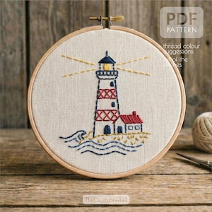 May include: Embroidered lighthouse design in a wooden hoop. The lighthouse is red and white with a blue top and is next to a small red-roofed building. The design includes blue waves and yellow light beams. The text "PDF PATTERN" is visible.