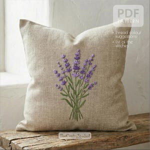 Peut inclure: Coussin en lin beige avec un bouquet de lavande brodé en violet et vert. Le coussin est posé sur une surface en bois rustique. Le texte sur le coussin indique "PDF PATTERN" et "+ suggestions de couleurs de fil + liste des points". TheCraftyStudio1 est également imprimé.