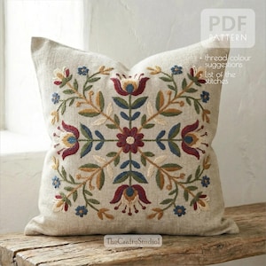 Può includere: Cuscino in lino beige con un ricamo floreale dettagliato. Il ricamo presenta un motivo simmetrico di fiori e foglie rosse, blu, gialle e verdi. Il cuscino è quadrato e poggia su una superficie di legno. Il testo sul cuscino recita "PDF PATTERN".