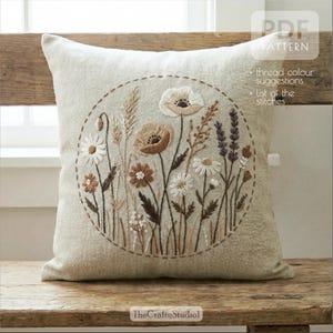Puede incluir: Cojín cuadrado beige con un diseño floral bordado. El diseño presenta una disposición circular de varias flores en tonos blancos, marrones y morados, con tallos y hojas verdes. El texto "PDF PATTERN" y "The Crafty Studio" son visibles.