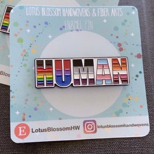 Puede incluir: Un pin de esmalte colorido con la palabra "HUMAN" en letras blancas. Cada letra está decorada con una bandera del orgullo diferente, incluidas las banderas del arcoíris, bisexual, transgénero y pansexual. El pin está sobre un fondo azul con estrellas blancas y el texto "LOTUS BLOSSOM HANDWOVENS & FIBER ARTS ENAMEL PIN".