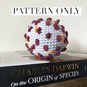 May include: Crochet pattern for a virus amigurumi, featuring a white base with maroon and orange accents. The finished product is a small, round ball with a textured surface.