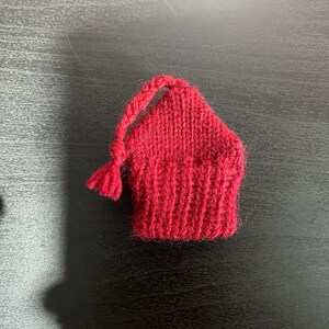 May include: A small, hand-knitted red hat with a tassel and a loop for hanging. The hat is made of a thick yarn and has a ribbed brim. The hat is likely a Christmas ornament or a small decorative item.