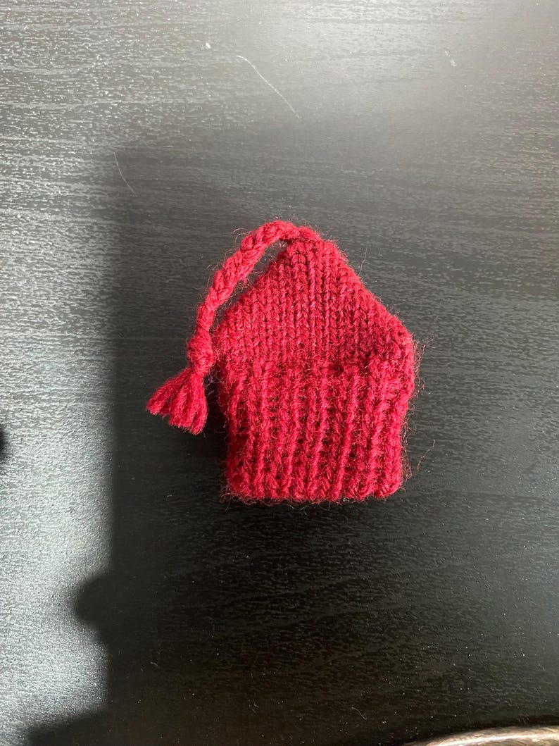 May include: A small, hand-knitted red hat with a tassel and a loop for hanging. The hat is made of a thick yarn and has a ribbed brim. The hat is likely a Christmas ornament or a small decorative item.