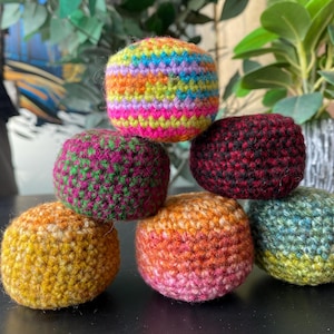 May include: A stack of six round, crocheted yarn balls in various colors. The top ball features a vibrant rainbow pattern, while the others display solid or variegated hues of yellow, orange, pink, green, and deep red, all with a textured stitch.