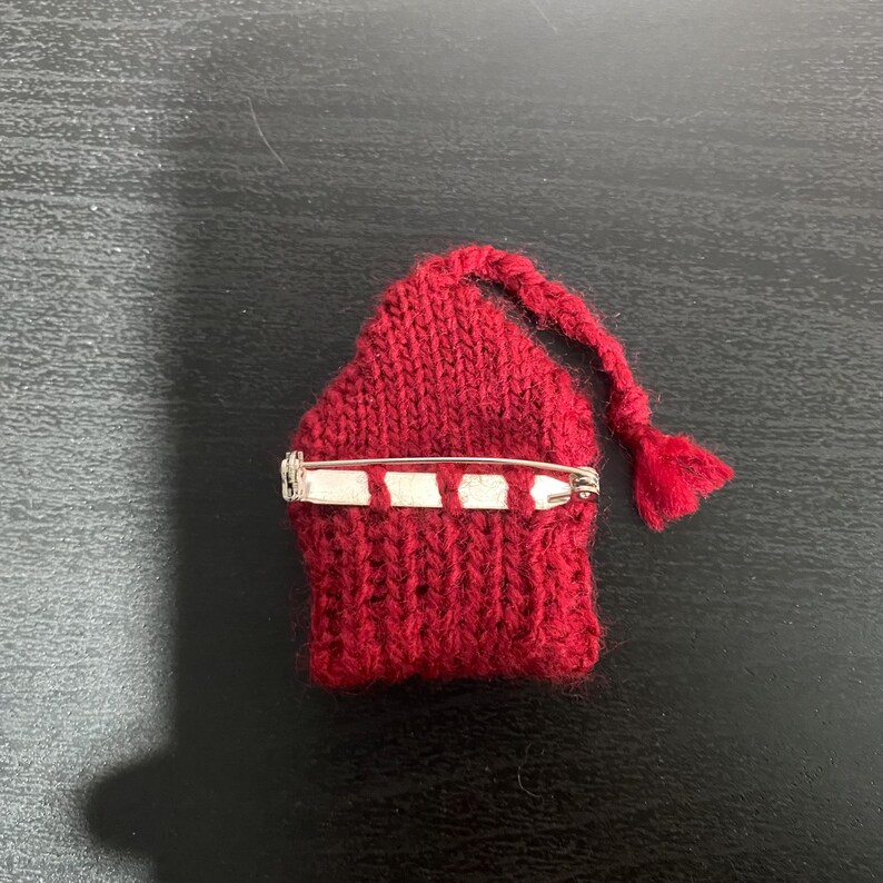 May include: A handmade, red knitted pin in the shape of a small house with a braided tassel. The pin has a silver safety pin clasp on the back. This unique accessory is perfect for adding a touch of whimsy to any outfit.