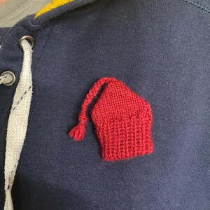 May include: A close-up of a navy blue hooded sweatshirt with a small, red, knitted hat-shaped patch. The sweatshirt has a yellow and gray collar and a white drawstring. The knitted patch is a vibrant red.